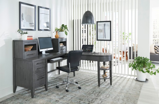 Modern home office featuring the aspenhome Preston Pedestal Desk and Return in a dark wood finish. The desk includes multiple storage drawers, a keyboard tray, and built-in shelving for organization. A black ergonomic chair complements the setup, while framed artwork, potted plants, and a neutral-toned rug add warmth. A vertical slatted room divider allows natural light to flow through, creating an open and airy workspace.